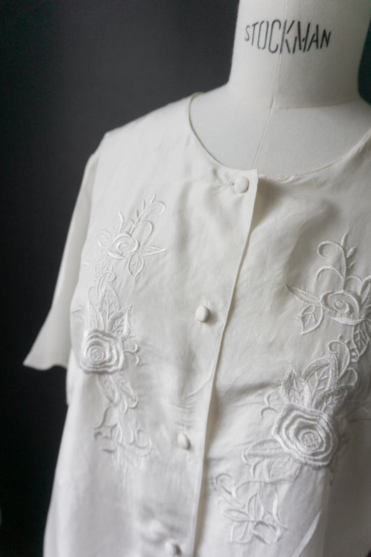 Weiße Vintage-Bluse mit floraler Stickerei und stoffbezogenen Knöpfen auf schwarzem Hintergrund